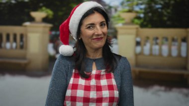 Parkta Noel Baba şapkası takan ve kırmızı önlüklü bir kadın yeşil ağaçların ve taş korkulukların arasında gülümsüyor; bayram neşesi şenliği ruhu.