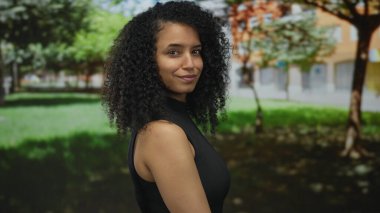 Genç bir kadın güneşli bir park ortamında tuğla bir binanın önünde çıplak omzunu ve çapraz kollarını gösteriyor..