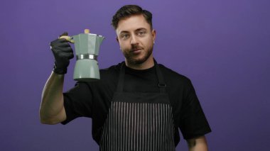 Önlüklü ve eldivenli genç beyaz adam mor arka planda moka demliği tutuyor, gülümseyerek kahve makinesini gösteriyor..