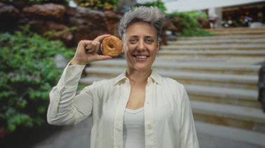 Kısa saçlı bir kadın binanın önünde yüzüne jöleli bir çörek tutarken gülümsüyor; sevinç tatmini.