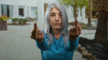 Yaşlı bir kadın, gri saçlı, orta parmaklı, açık hava ortamında mavi bluz giyiyor..