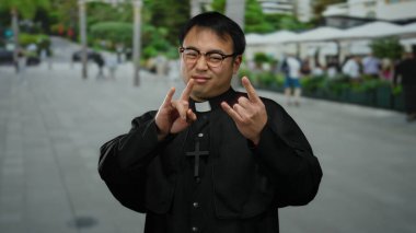 Gözlüklü bir rahip, şehir dışında, arka planda insanlar ve yeşillik olan bir yerde jest yapıyor..