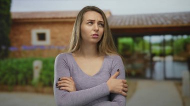 Dışarıda genç bir kadın, yeşil ve mavi çiçeklerle çevrili kırsal bir binanın önünde kolları çaprazlanmış sıradan bir kıyafet giyiyor..