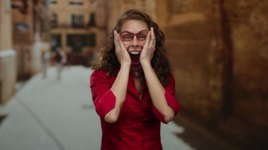 Kırmızı gözlüklü ve kırmızı gömlekli bir kadın sıcak, güneşli bir ortamda güvenle gülümsüyor..