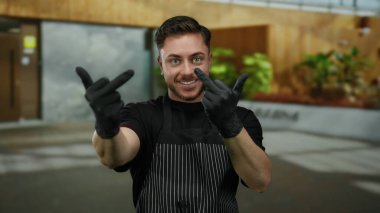 Sakallı genç adam orta parmaklarını şehir sokaklarında gösteriyor. Açık havada eldiven ve önlük giyiyor. Gülümseyerek ve gelişigüzel bir tavırla meydan okuyor..