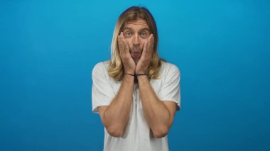 Uzun saçlı, beyaz tişörtlü sarışın bir adam parlak mavi stüdyo ortamında iki elini de geniş gözlü yanaklara bastırıyor..
