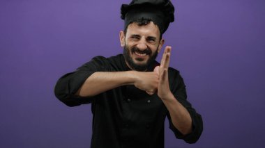Siyah üniformalı İspanyol erkek şef, mor bir arka plana karşı el çırpıyor, neşeli ve enerjik bir ifade sergiliyor..