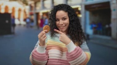 Canlı bir caddedeki kurabiyeye gülümseyen bir kadın renkli bir kazakla dışarıda mutluluk ve neşe sergiliyor canlı bir şehir ortamında eğlenceli bir anı vurguluyor..