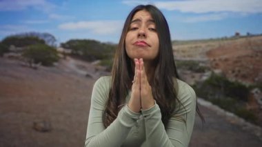 Açık hava parkında dua eden elleri olan genç bir kadın engin mavi gökyüzünün altında manzaralı bir arka plana karşı umut dolu bir ifade sergiliyor..