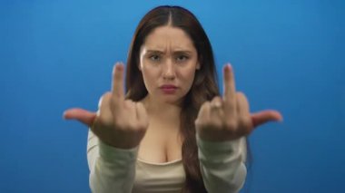 Kadın, parlak mavi bir stüdyo sahnesinde orta parmaklarını kaldırır çatık kaşlı ve büzüşmüş dudaklarıyla; meydan okuma.