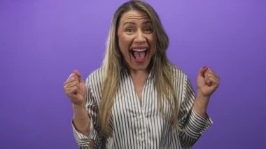 Orta yaşlı sarışın kadın çizgili gömlek giyiyor. Yumruk kaldırıyor ve mor stüdyoda gülümsüyor. Heyecan verici bir coşku..