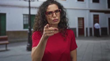Kırmızı gömlekli ve kıvırcık saçlı gözlüklü bir kadın şehir caddesinde kamerayı işaret ediyor..