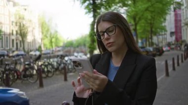 Amsterdam Kanalları yakınlarında akıllı telefon kullanan, gözlüklü, açık şehir ortamında bisikletlerle çevrili, yeşil ağaçlar ve park halindeki arabalarla çevrili bir kadın.