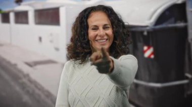 Dışarıdan gülümseyen, kıvırcık saçlı, kazak giyen, çöp kutusunun yanına işaret eden bir kadın, kentsel geri dönüşüm sahnesinde mutluluğu öneriyor..