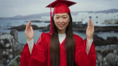 Kırmızı mezuniyet elbisesi giymiş bir kadın bulutlu gökyüzünün altında deniz kenarında gezinti yapıyor.