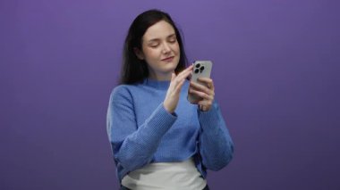 Mor arka plan önünde elinde akıllı telefonla gülümseyen bir kadın mavi kazak giyiyor meşgul görünüyor ve teknolojiye hazır dijital yaşam tarzıyla iletişim kuruyor..