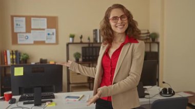 Ofiste gülümseyen bir kadın gözlük takıyor ve bej ceket giyiyor. Çalışmalarını bilgisayar ve kitaplarla sunuyor. Belgeler ve bitkilerle dolu bir iş ortamı öneriyor..