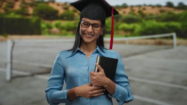 Tenis kortunda güvenle gülümseyen bir kadın mezuniyeti kutlamak için kep cüppeli bir kitapla dışarıda başarıyı ve başarıyı ifade eden mutlu Latin bir öğrenci olarak.