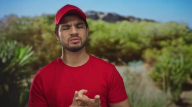 Kırmızı tişörtlü ve kepli bir adam ormanda parmaklarını sayıyor yoğun yeşil ağaçlarla; odaklanmış planlama.