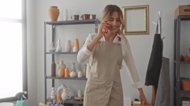 Çömlek atölyesinde telefonla konuşan genç bir kadın siparişleri kontrol ederken ve kil aletlere uzanırken; yaratıcılık odaklı sükunet.