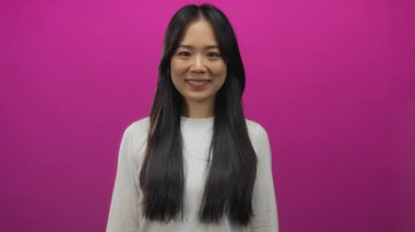 Canlı pembe bir arka plan önünde gülümseyen uzun siyah saçlı beyaz bir gömlek giyen ve kendine güvenerek kendini işaret eden bir kadın..