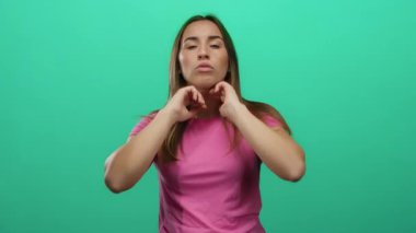 Pembe gömlekli bir kadın, izole edilmiş yeşil bir arka planın önünde kendini yelpazeliyor, sıcaklık ve rahatsızlığı ifade ediyor, jestleri serinleme ihtiyacını gösteriyor, sıcak ve telaşlı görünüyor..