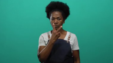 Önlüklü kadın tetikte ve yeşil arka plana karşı tehdit oluşturuyor. İfadeler ve el işaretleriyle yoğun bir odaklanma ile iddialı iletişim kuruyor..