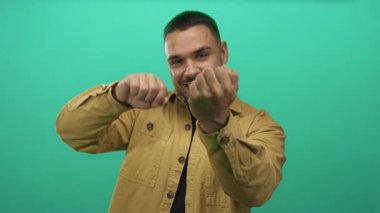 Yeşil stüdyoda orta parmak hareketi yapan adam geniş sırıtıyor ve öne doğru eğiliyor..