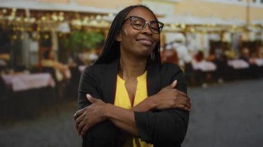 Açık hava pazarında kollarını kavuşturmuş gülümseyen bir kadın, şehir atmosferini sıcak bir ifadeyle harmanlıyor, gözlük takıyor ve sıradan bir kıyafet giyiyor.