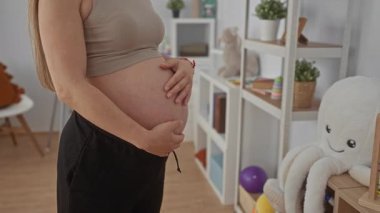 Bebek odasında karnını oyuncaklarla ve raflarla sarmış hamile bir kadın annelik için huzurlu ve umut verici bir atmosfer yaratıyor..