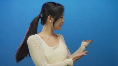 Genç bir kadın, her iki elini de hafifçe yana uzatarak parlak mavi bir arka plana karşı sunum yapıyor, neşeli ifadesini ve davetkar hareketini vurguluyor..