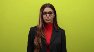 Gözlüklü ve takım elbiseli, sarı renkli bir arkaplan karşısında tek başına duran bir kadın.