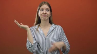 Sarı saçlı, çizgili tişörtlü bir kadın iki elini de stüdyoda tutarak turuncu arka planda belirsizlik yaratıyor..