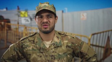 Kamuflaj üniformalı adam bina yakınında eskrim yaparken yüzünü buruşturuyor; görev basıncının düşmesi.