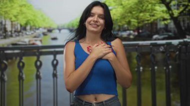 Latin kökenli genç bir kadın sıcak öğleden sonra ışığında şehir caddesinde kanalın korkuluklarında çıplak ellerini göğsüne koyuyor..