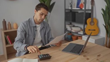 Adam kağıt üzerine not yazıyor, tahta bir masada dizüstü bilgisayarın yanında sarı kalemle ve stüdyoda hesap makinesiyle; verimliliğe odaklanıyor..