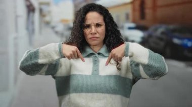 Kıvırcık saçlı ve gözlüklü bir kadın şehir merkezinde kendine güvenen çizgili bir süveter giyiyor..