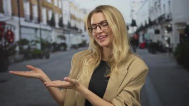 Şehir caddesinde sarışın ve gözlüklü bir kadın gündüz vakti iki eliyle bej ceket giyiyor.