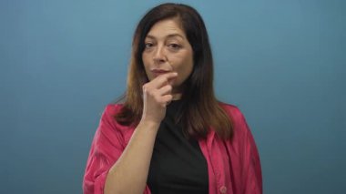 Pembe ceketli orta yaşlı İspanyol kadın stüdyoda sessizlik için parmaklarını dudaklarına bastırıyor..