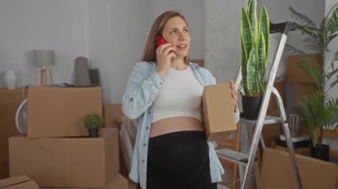 Yeni ev oturma odasında telefon kulübesinde konuşan hamile bir kadın. Etrafı kutu ve bitkilerle çevrili..