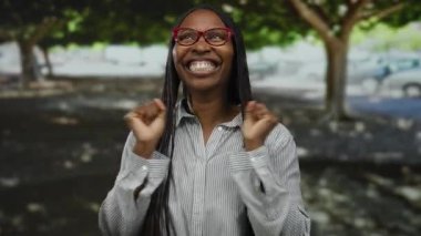 Canlı bir parkta gülümseyen, ağaçların arasından süzülen, neşeli ifadesini vurgulayan, gözlük takan, çizgili tişörtlü, mutluluk ve güven saçan bir kadın..