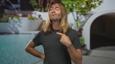 Koyu gri tişörtlü, uzun sarı saçlı bir adam havuz kenarındaki bina girişinde parmağıyla göğsünü işaret ediyor..