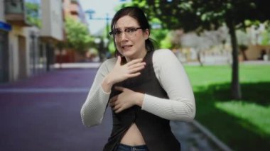 Dışarıdaki gözlüklü esmer kadın sessiz bir sokakta korkmuş görünüyor. Endişeli bir ifadeyle göğsüne sarılıyor. Huzurlu bir şehir ortamındaki korkuyu işaret ediyor..