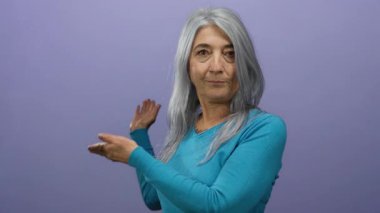 Deniz mavisi süveterli gri saçlı yaşlı kadın, boş bir mor arka plana doğru jest yapıyor, kopya metni için bir alan yaratıyor ve yön ve sunum kavramlarını destekliyor..