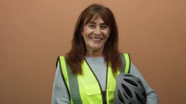 Senior hispanic woman smiling in a brown background wearing a reflective vest and holding a helmet, showcasing safety and readiness.