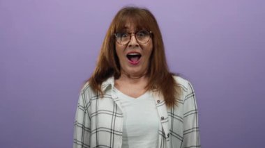 Şaşkın bir ifade ve gözlüklü İspanyol son sınıf öğrencisi kadın canlı bir mor arka plana karşı duruyor, elleri yüzü üzerinde, ekose bir gömlek giyiyor..