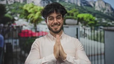 Genç, sakallı, gözlüklü, dua ederken gülümseyen, yeşil ve dağlarla çevrili genç bir İspanyol.