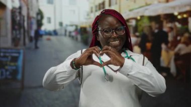 Woman with stethoscope smiling and forming heart shape with hands on city street terrace with outdoor restaurant.