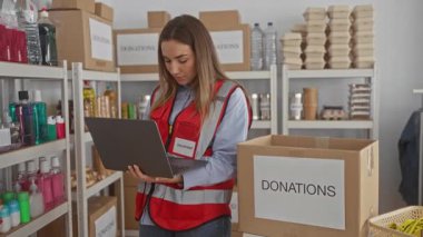 İç mekândaki hayır kurumlarında dizüstü bilgisayar kullanan genç bir kadın. Etrafı kutularla ve malzeme raflarıyla çevrili. Kırmızı yelek giymiş, insani yardım organizasyonları düzenliyor.