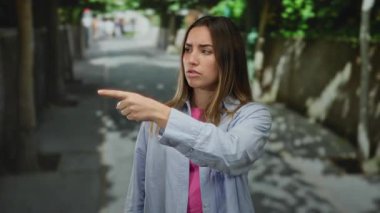 Ağaç kaplı bir caddeyi işaret eden kadın, sıradan bir gömlek giyiyor ve şehir ortamında ciddi görünüyor, odak ve yön anını yakalıyor..